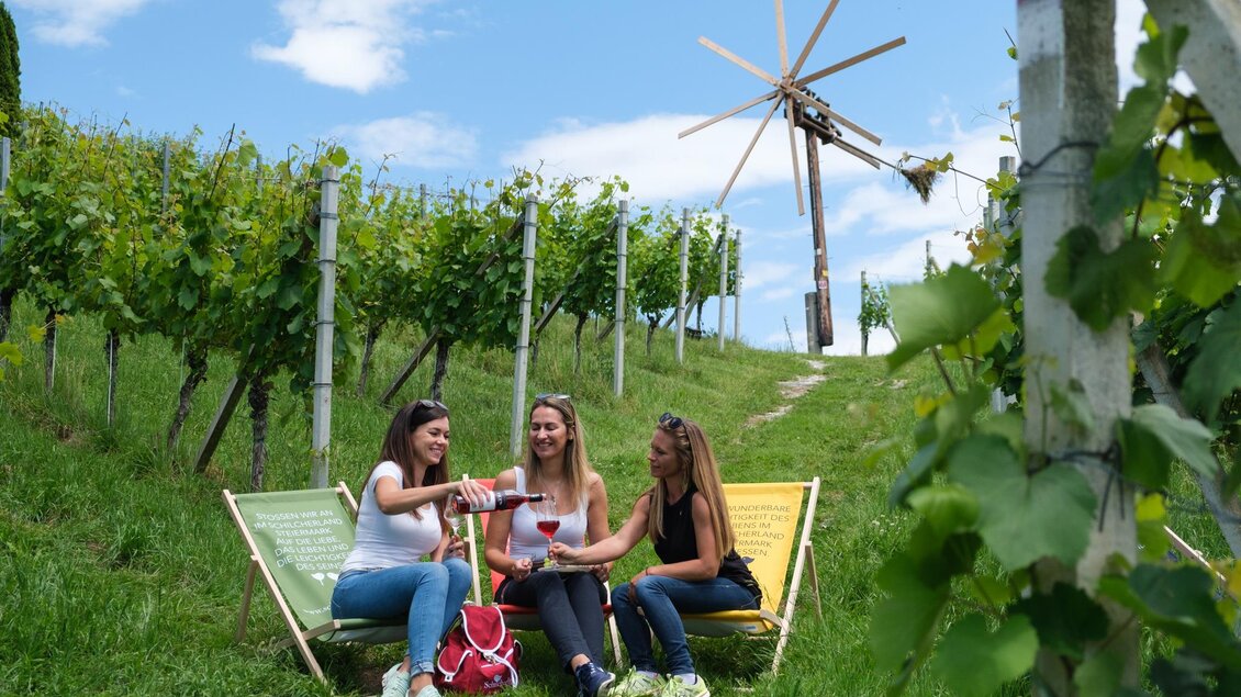 Drei Frauen entspannen in Liegestühlen zwischen Weinreben. Im Hintergrund steht ein Windrad unter einem blauen Himmel. | © Schilcherland Steiermark