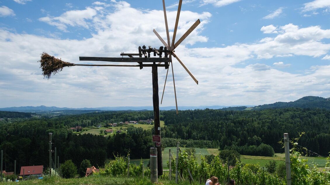 Eine ländliche Landschaft mit einer Windmühle und strahlend blauem Himmel. Im Vordergrund sitzen Menschen auf einer Wiese und genießen die Aussicht. | © Schilcherland Steiermark