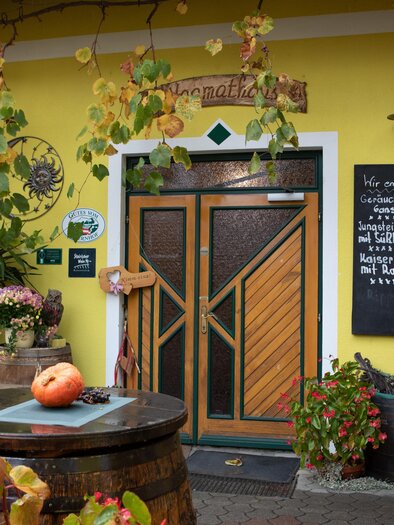 Ein einladendes, gelbes Gebäude mit Holzverzierungen und einer Eingangstür. Vor der Tür stehen Pflanzen und eine Tafel mit Schrift. | © GlaMUR-Genuss am Fluss