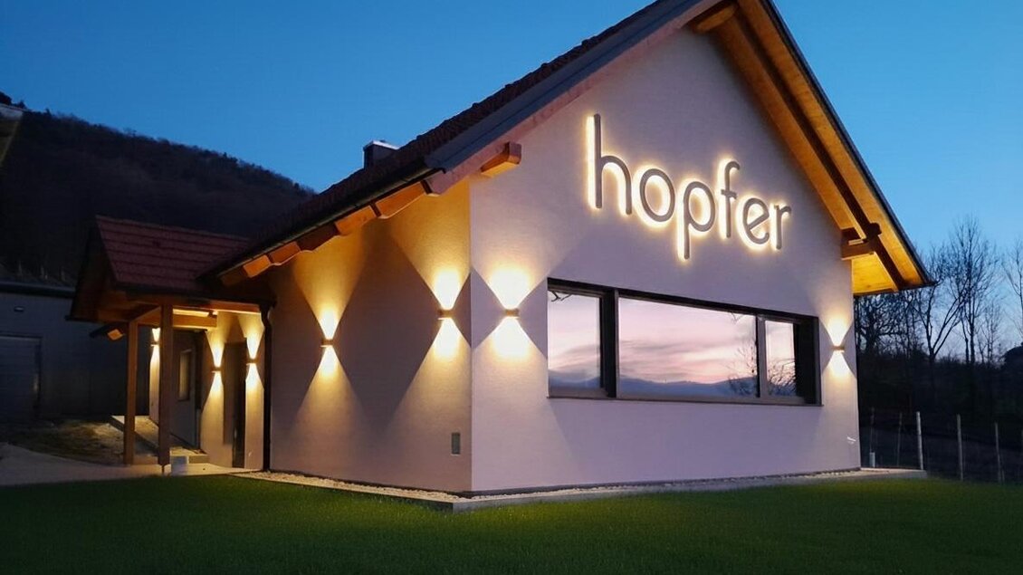 Ein modernes Gebäude mit der Schrift "hopfer" an der Fassade. Die Außenbeleuchtung sorgt für eine einladende Atmosphäre in der Dämmerung.