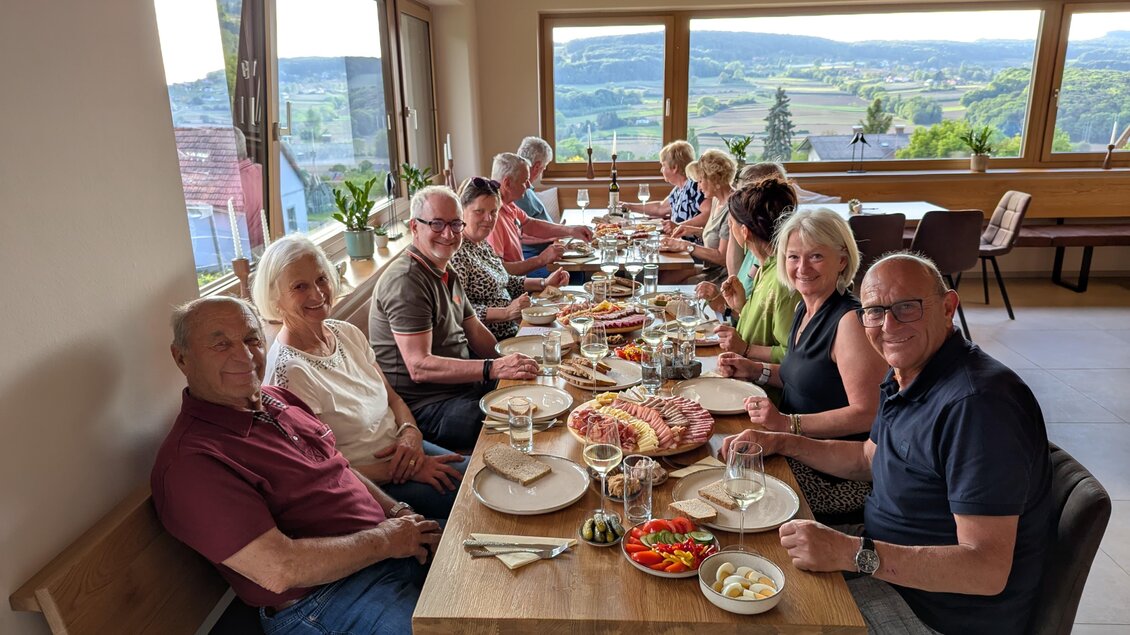 Eine Gruppe von Menschen sitzt an einem langen Tisch und genießt eine Mahlzeit. Im Hintergrund sind große Fenster mit Blick auf die Landschaft.