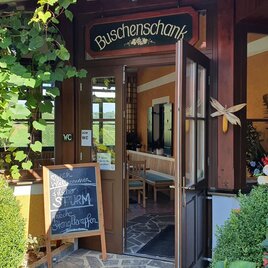 Ein einladender Eingang zu einem Buschenschank mit einer gemütlichen Atmosphäre. Umgeben von Pflanzen und einer Tafel mit Angeboten. | © Weingut Ilgi