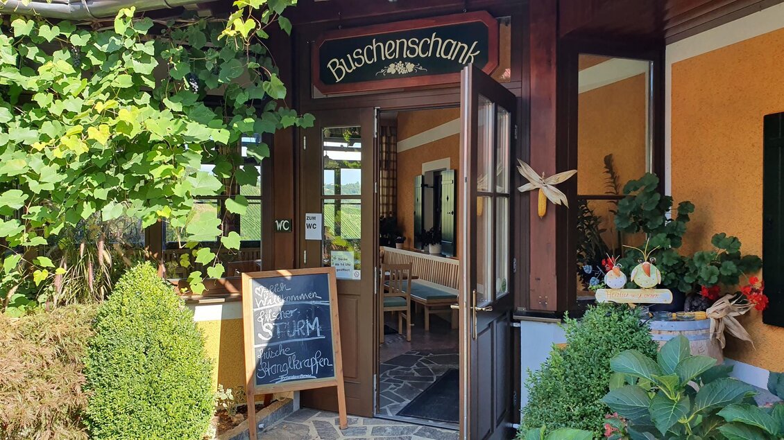 Ein einladender Eingang zu einem Buschenschank mit einer gemütlichen Atmosphäre. Umgeben von Pflanzen und einer Tafel mit Angeboten. | © Weingut Ilgi