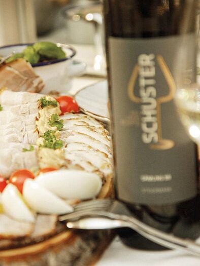 Ein appetitliches Anrichten von Fleisch und Eiern auf einem Holzbrett, umgeben von frischen Tomaten und Kräutern. Im Hintergrund steht eine Flasche Wein und ein Glas. | © Klöcklschuster