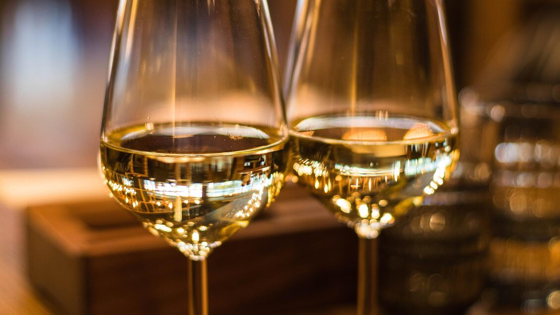 Zwei elegante Weingläser mit weißem Wein stehen auf einem Tisch. Im Hintergrund sind unscharfe Töne von Holz und Glas zu sehen. | © Weingut Luttenberger