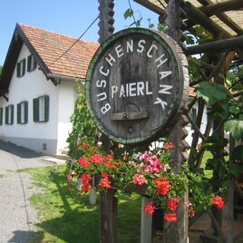 Ein rustikales Schild mit der Aufschrift „Buschenschank Paierl“ hängt an einer Holzkonstruktion. Im Hintergrund sieht man ein gemütliches Gebäude mit grünen Fensterläden. | © Buschenschank Paierl