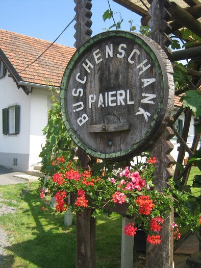 Ein rustikales Schild mit der Aufschrift „Buschenschank Paierl“ hängt an einer Holzkonstruktion. Im Hintergrund sieht man ein gemütliches Gebäude mit grünen Fensterläden. | © Buschenschank Paierl