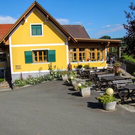 Ein freundliches, gelb gestrichenes Haus mit grünen Fensterläden. Draußen sind gemütliche Sitzplätze mit Tischen und Stühlen angeordnet. | © Rack-Buschenschank