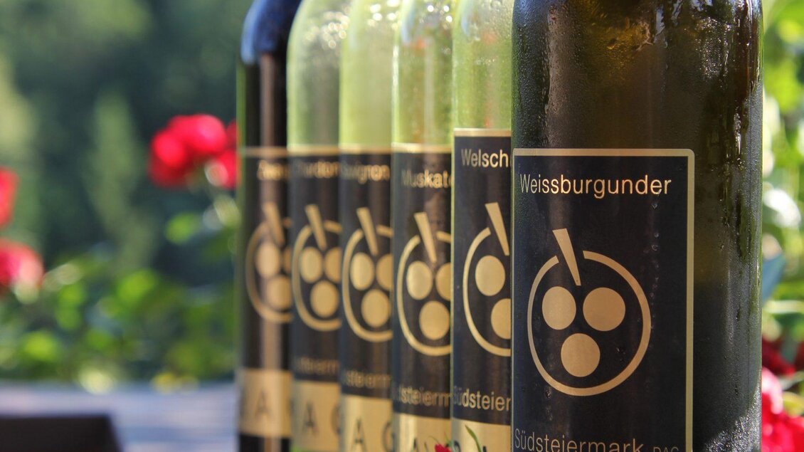 Mehrere Flaschen Wein mit Etiketten stehen auf einem Tisch, umgeben von roten Rosen. Im Hintergrund sind grüne Bäume und eine ruhige Landschaft zu sehen. | © Rack-Buschenschank