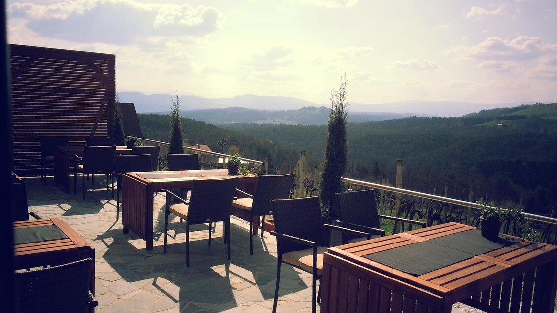 Eine Terrasse mit Holztischen und Stühlen bietet einen weiten Blick auf die Landschaft. Die Szenerie ist von sanften Hügeln und blauem Himmel umgeben. | © Weingut Reiterer