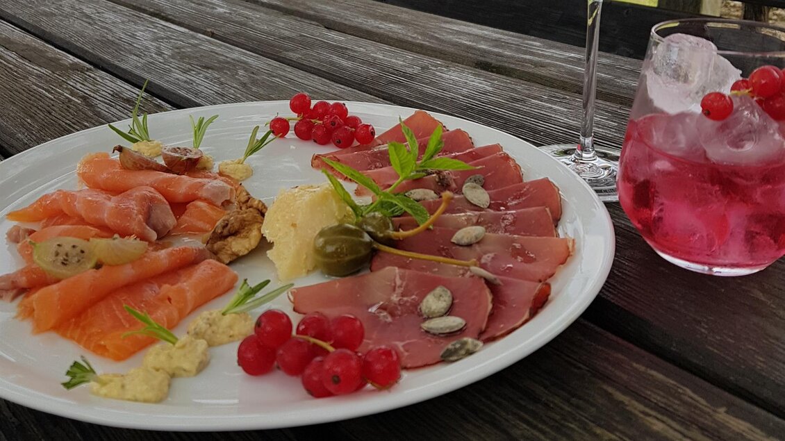 Ein Teller mit verschiedenen Fisch- und Fleischsorten, garniert mit Kräutern und roten Johannisbeeren. Daneben steht ein Glas mit einem fruchtigen Getränk und Eiswürfeln. | © Weingut Reiterer