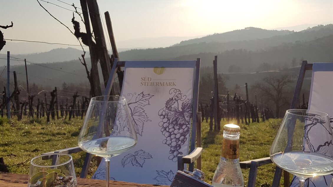 Eine idyllische Weinszene mit Gläsern und einer Flasche auf einem Holztisch. Im Hintergrund sind sanfte Hügel und die Sonne über den Weinbergen zu sehen. | © Weingut Reiterer