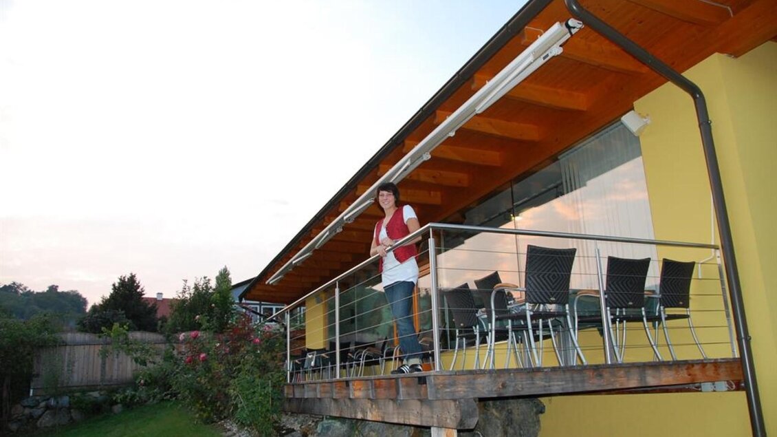 Eine Person steht auf dem Balkon eines modernen Hauses. Der Bereich ist mit Stühlen und Tischen ausgestattet und umgeben von einer grünen Gartenlandschaft. | © Weingut &  Buschenschank Stefanie Sauer