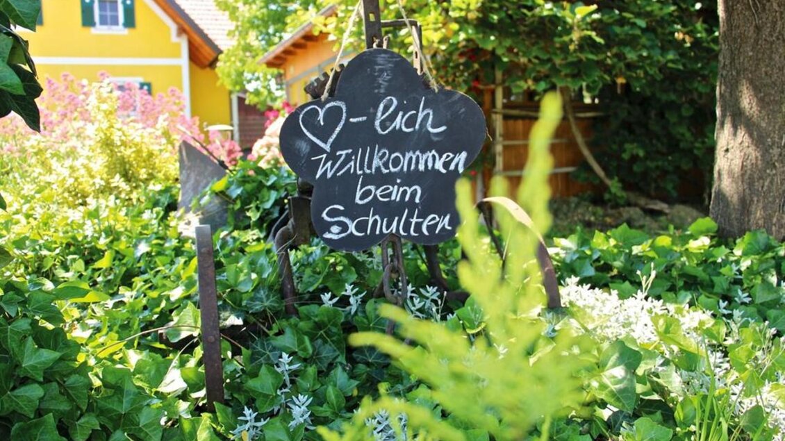 Eine einladende Tafel steht in einem grünen Garten. Sie heißt die Besucher mit „Willkommen beim Schütten“ willkommen. | ©  Buschenschank Schulter