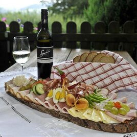Ein schön angerichteter Brett mit verschiedenen Wurst- und Käsesorten, begleitet von frischem Gemüse. Dazu stehen ein Glas Weißwein und eine Flasche auf einem Tisch mit grüner Landschaft im Hintergrund. | © Buschenschank Stoff