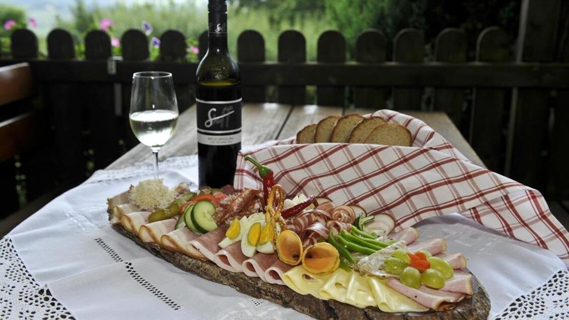 Ein schön angerichteter Brett mit verschiedenen Wurst- und Käsesorten, begleitet von frischem Gemüse. Dazu stehen ein Glas Weißwein und eine Flasche auf einem Tisch mit grüner Landschaft im Hintergrund. | © Buschenschank Stoff