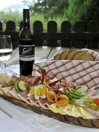 Ein schön angerichteter Brett mit verschiedenen Wurst- und Käsesorten, begleitet von frischem Gemüse. Dazu stehen ein Glas Weißwein und eine Flasche auf einem Tisch mit grüner Landschaft im Hintergrund. | © Buschenschank Stoff