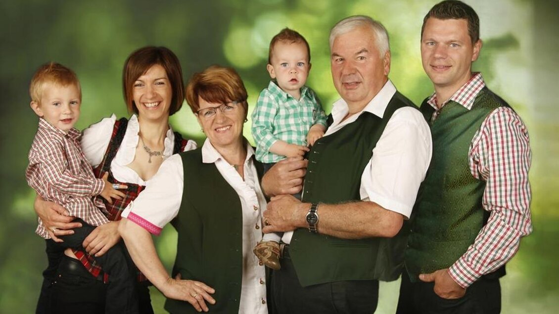 Eine fröhliche Familie posiert gemeinsam in traditioneller Kleidung. Sie stehen vor einem unscharfen, grünen Hintergrund und lächeln. | © Buschenschank Stoff