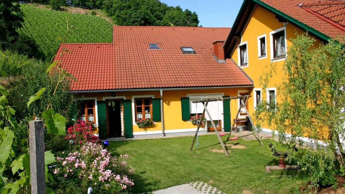 Ein charmantes Haus mit gelber Fassade und roten Dächern. Der Garten ist grün und blühend, ideal für entspannte Tage im Freien. | © Buschenschank Stoff