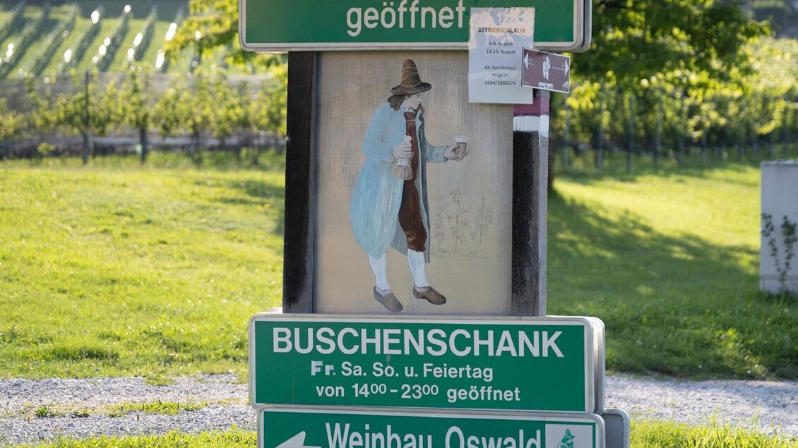 Ein Wegweiser mit Informationen zu einem Flaschenweinsverkauf und einem Buschenschank. Im Hintergrund sieht man Weinberge und eine grüne Wiese. | © TV Südsteiermark - Dominik Feibel