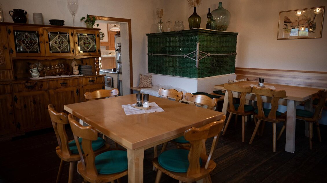 Ein einladendes Restaurant mit Holztischen und Stühlen. Im Hintergrund steht ein grüner Kachelofen und eine Holzvitrine an der Wand. | © TV Südsteiermark - Dominik Feibel