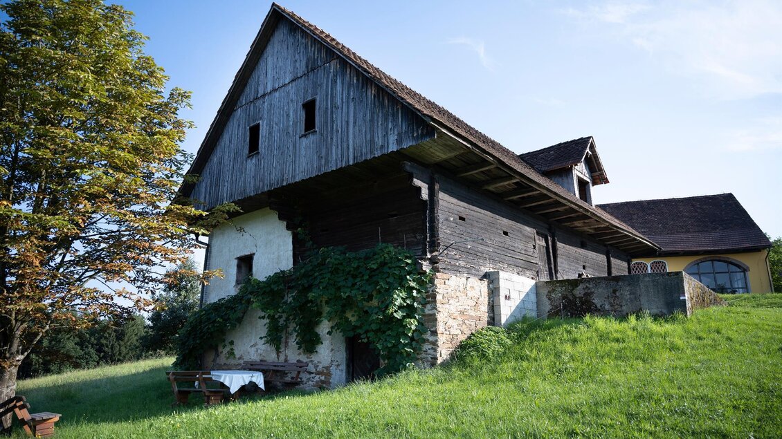 Ein rustikales Bauernhaus mit einer Holzfassade steht auf einer Wiese. Der Himmel ist klar und ein Baum ist in der Nähe zu sehen. | © TV Südsteiermark - Dominik Feibel