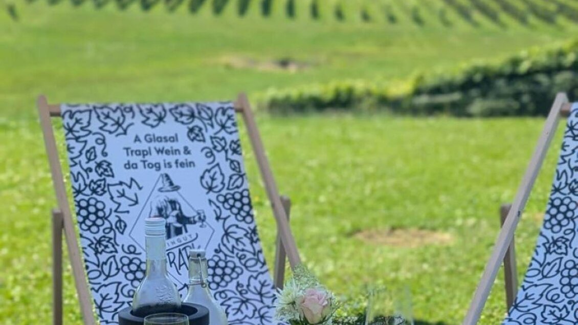 Zwei Liegestühle stehen auf einer grünen Wiese mit Weinreben im Hintergrund. Auf einem Holzkasten sind Gläser mit Wein und Wasser sowie eine kleine Blume platziert. | © Andrea Bayr