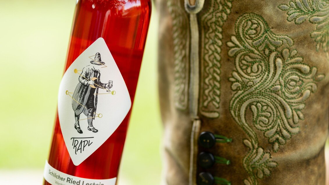Ein Mann in traditionellen Lederhosen hält eine Flasche mit rosafarbenem Getränk in der Hand. Die Flasche zeigt ein kreatives Etikett mit einer lustigen Figur. | © Trapl - Nikola Milatovic