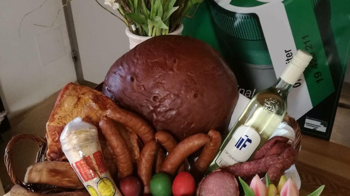 Ein Osterschinkenkorb mit verschiedenen Wurstsorten, bunten Eiern und einem Flasche Wein. Die Dekoration enthält frische Blumen und ein Festtagslogo im Hintergrund. | © Weinhof Tropper