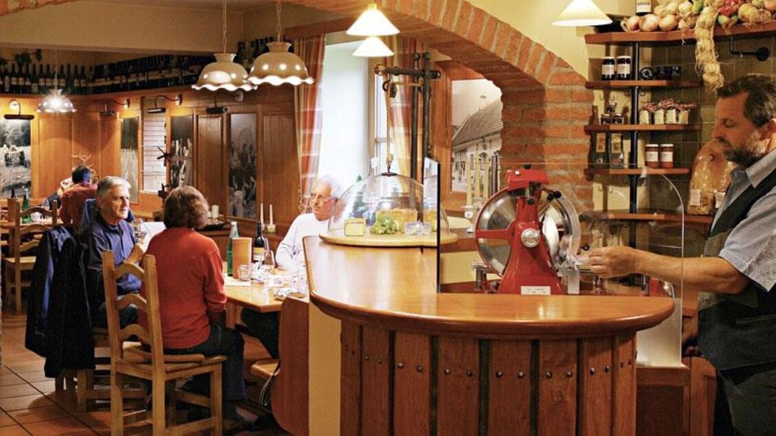 Ein gemütliches Restaurant mit Holzmöbeln und warmem Licht. Gäste genießen ihr Essen, während ein Kellner an der Theke arbeitet. | © Weingut Trummer