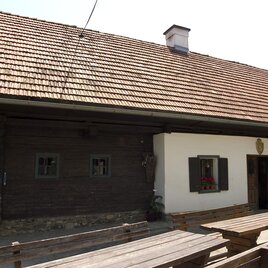 Ein traditionelles Haus mit einem roten Ziegeldach und Holzverkleidung. Vor dem Haus stehen Holztische und Stühle. | © Familie Schilcher | Weingut Wölfl