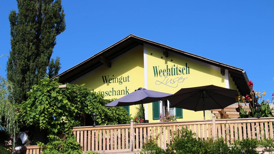 Ein sonniges Gebäude mit gelber Fassade und einer Holzterrasse. Es hat einen Sonnenschirm und ist von Pflanzen umgeben. | © Weingut Wechtitsch