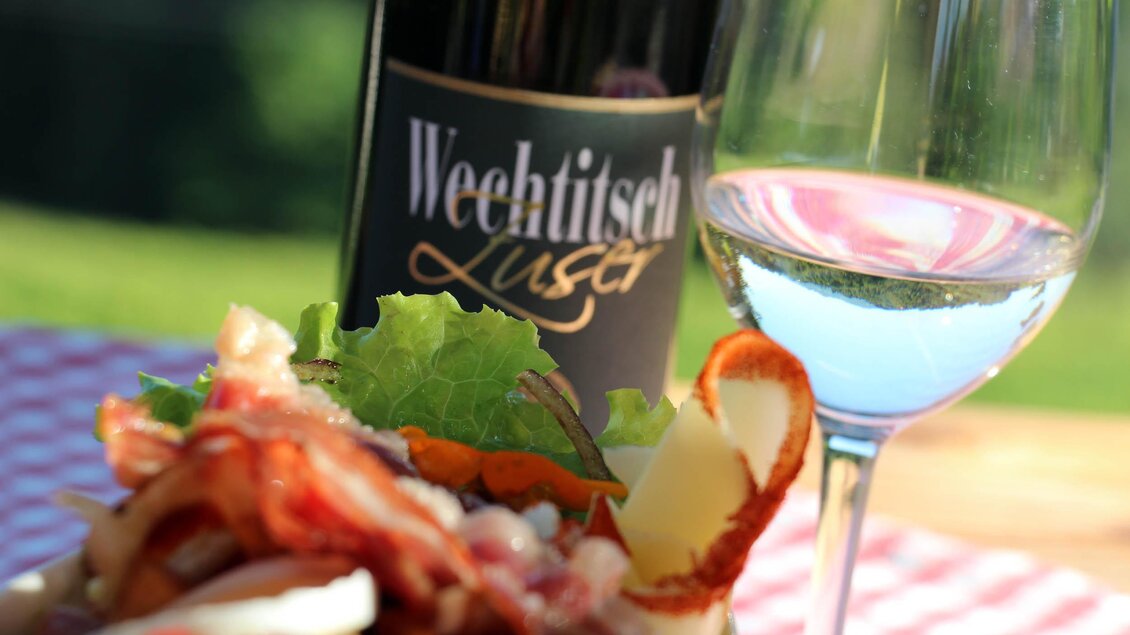 Ein gedeckter Tisch mit einem köstlichen Salat, Speck und frischem Gemüse. Im Hintergrund steht eine Flasche Wein und ein Glas mit klarer Flüssigkeit. | © Weingut Wechtitsch