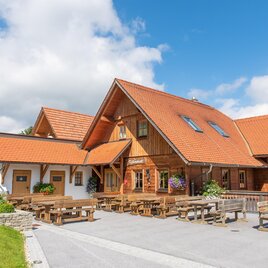 Ein traditionelles Holzhaus mit orangefarbenem Dach und gemütlicher Terrasse. Die Umgebung ist grün und einladend, ideal für ein sonniges Wochenende. | © Buschenschank Kellerstöckl