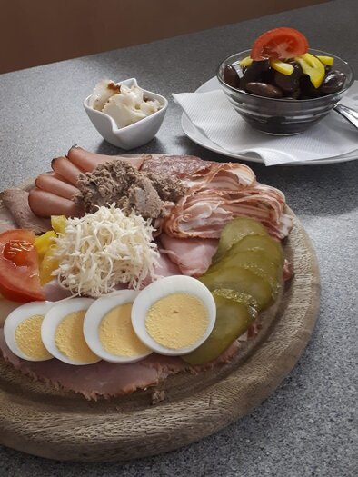 An appetizing plate with various sausage and cheese variations, eggs, and vegetables. In the background, a dessert made of chocolate and fruit. | © Buschenschank Wiesenhofer