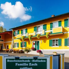Ein hellgelbes Gebäude mit grünen Fensterläden und einem einladenden Außensitzbereich. Das Schild weist auf den Buscheneschank und Hofladen der Familie Zach hin. | © TV Region Graz - Harry Schiffer