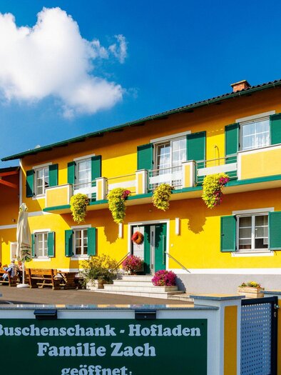 A light yellow building with green shutters and an inviting outdoor seating area. The sign indicates the family Zach's bush tavern and farm shop. | © TV Region Graz - Harry Schiffer