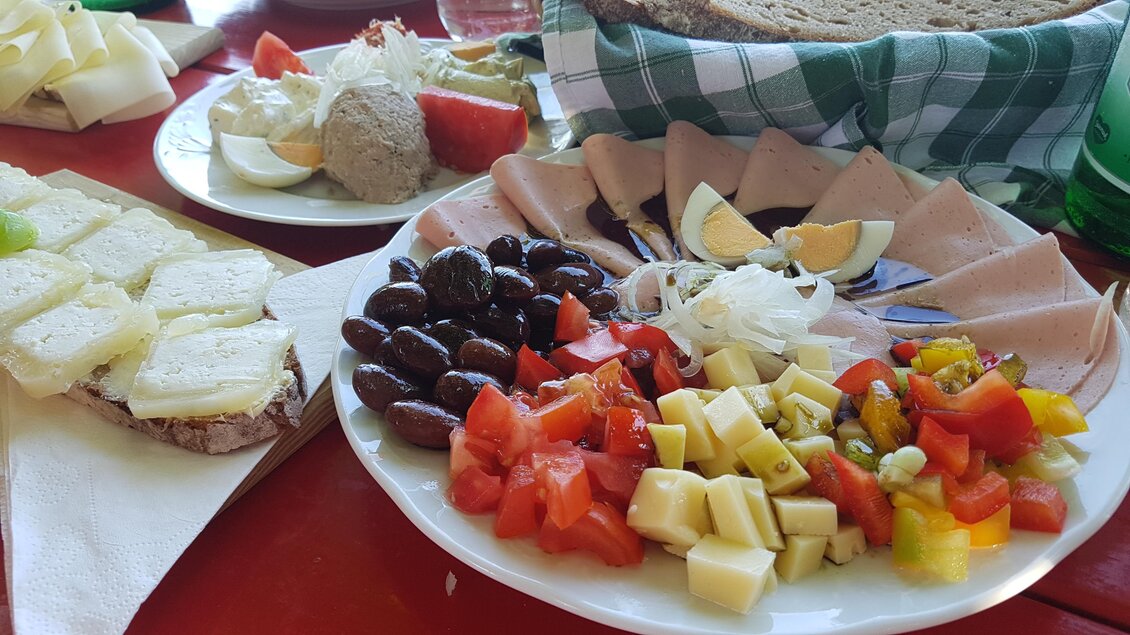 Ein Teller mit verschiedenen Käsesorten, Oliven, Tomaten und einer Scheibe Wurst. Im Hintergrund sind weitere Snackplatten und Brot sichtbar. | © Tourismusverband Oststeiermark