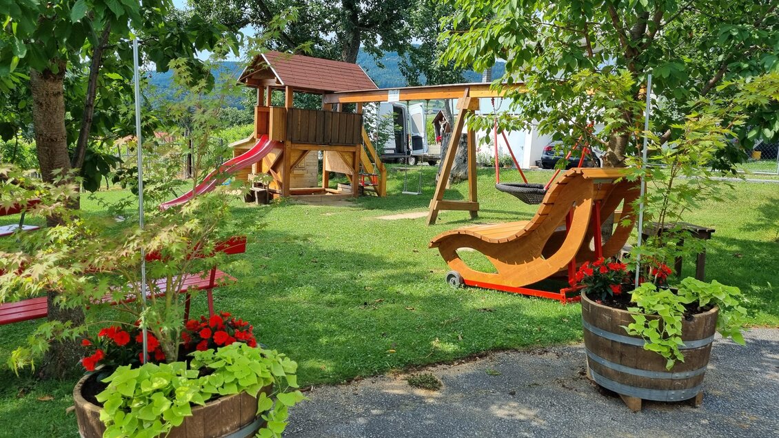Ein schöner Spielplatz mit einer Rutsche, Schaukeln und einem Holzspielhaus. Bunte Pflanzen in Holztöpfen umgeben die gepflegte Grasfläche. | © Schweighofer