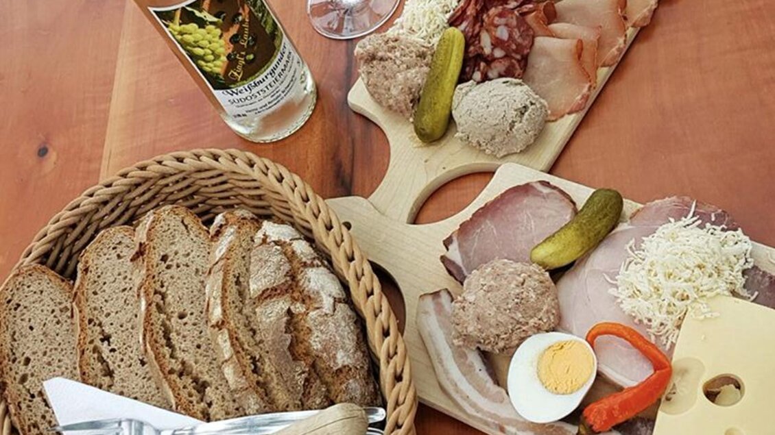 Ein rustikales Brett mit Wurst, Käse, Eiern und eingelegtem Gemüse. Dazu frisches Brot in einem Korb und ein Glas Weißwein. | © Buschenschank Ziagls Laube