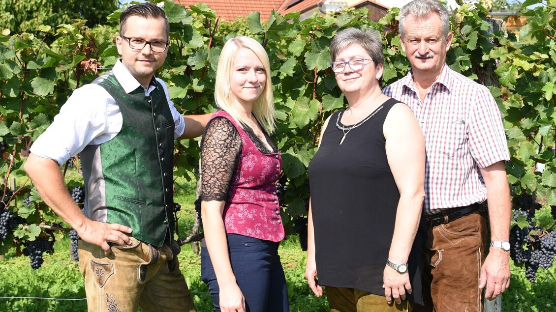 Eine Familie steht in einem Weinfeld. Sie tragen traditionelle Trachten und lächeln zur Kamera. | © Buschenschank Ziagls Laube