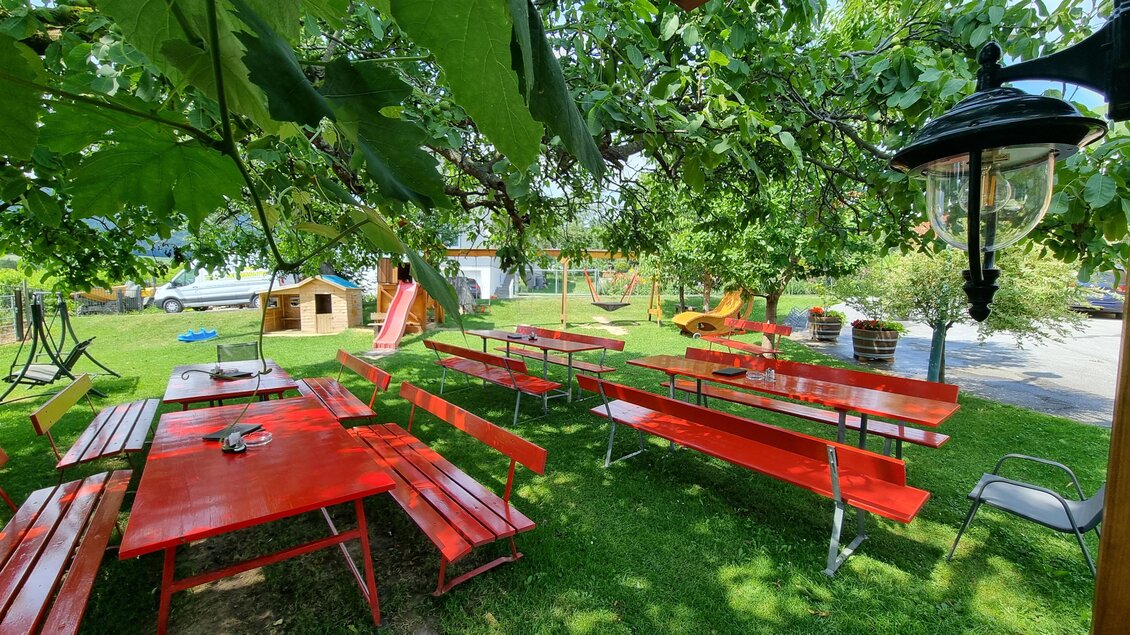 Ein einladender Garten mit roten Tischen und Bänken unter grünen Bäumen. Im Hintergrund sind Spielgeräte und ein kleines Häuschen zu sehen. | © Schweighofer