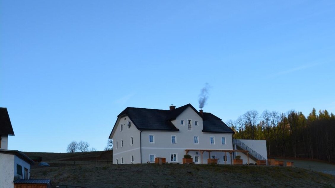 Ein großes, weißes Haus steht auf einem Hügel unter einem blauen Himmel. Rauch steigt aus dem Schornstein auf und die Umgebung ist ruhig und ländlich. | © Bichler