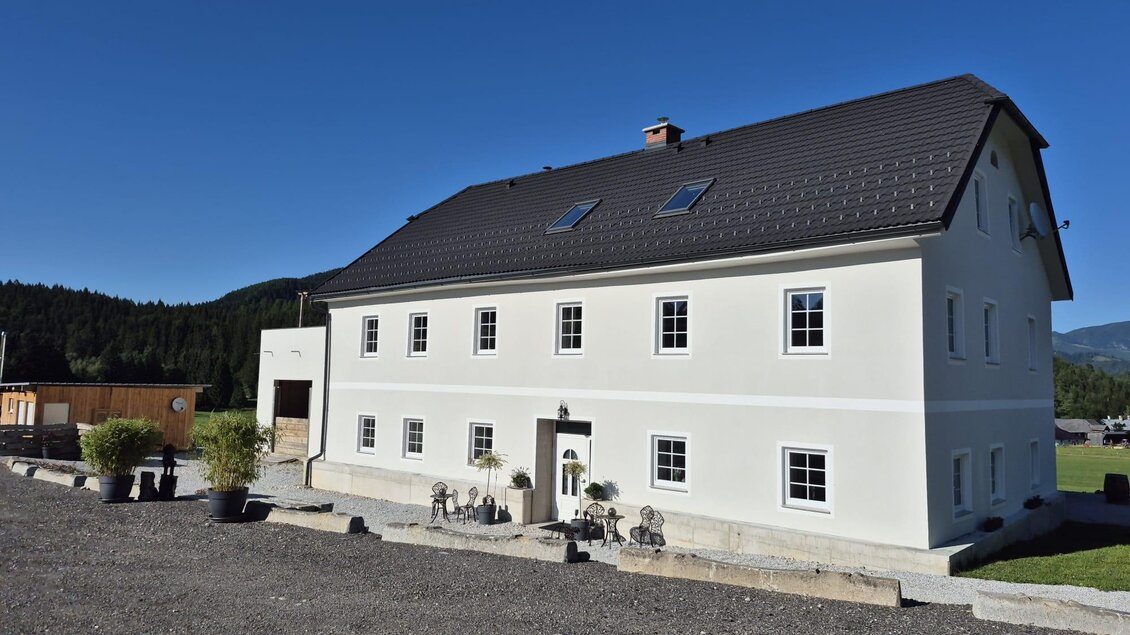Ein modernes, zweigeschossiges Haus mit weißer Fassade und schwarzen Dach. Die Umgebung ist ländlich und ruhig, umgeben von Bäumen und Grünflächen. | © TV Hochsteiermark