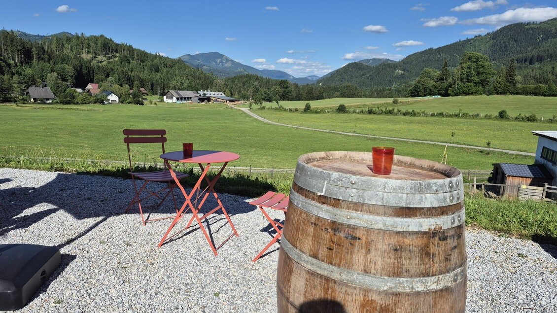Eine idyllische Landschaft mit einem Blick auf die Berge. Im Vordergrund stehen ein Holzfass und ein roter Tisch mit Stühlen. | © TV Hochsteiermark