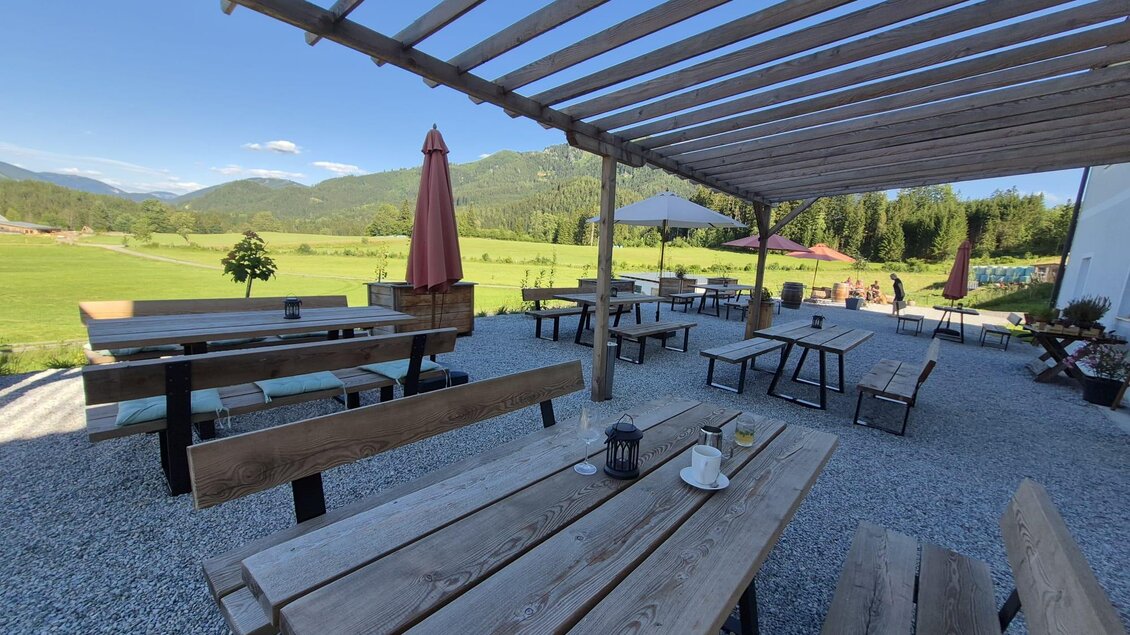 Eine gemütliche Außenterrasse mit Holztischen und Bänken. Im Hintergrund sind grüne Wiesen und Berge sichtbar. | © TV Hochsteiermark