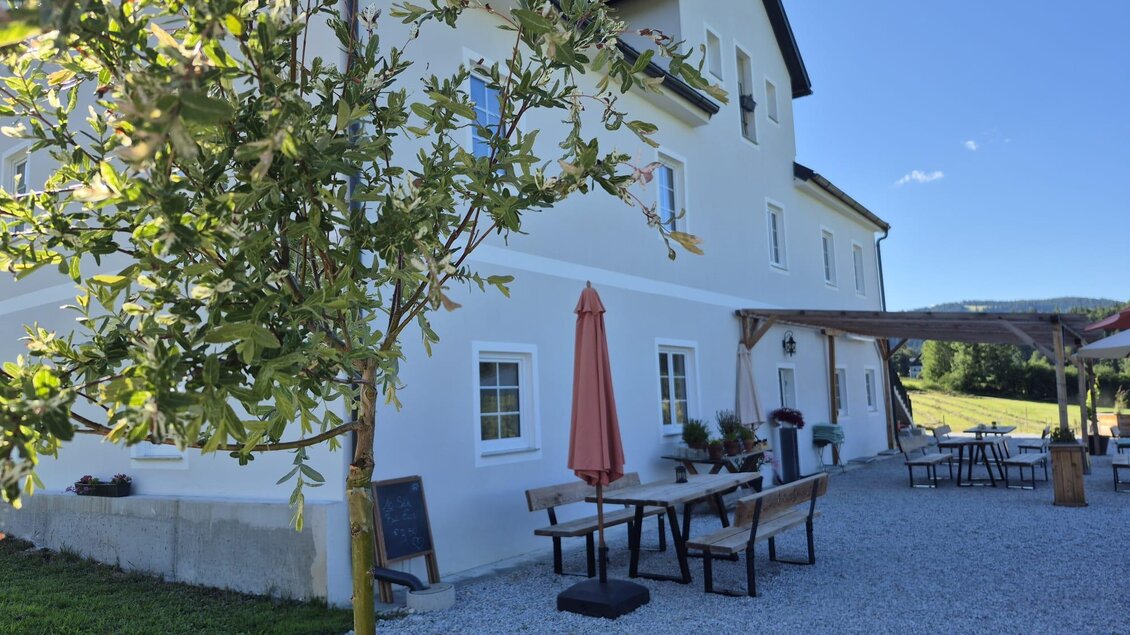 Ein modernes Gebäude in ländlicher Umgebung mit einem Innenhof. Es gibt Tische, Stühle und einen Sonnenschirm unter blauem Himmel. | © TV Hochsteiermark