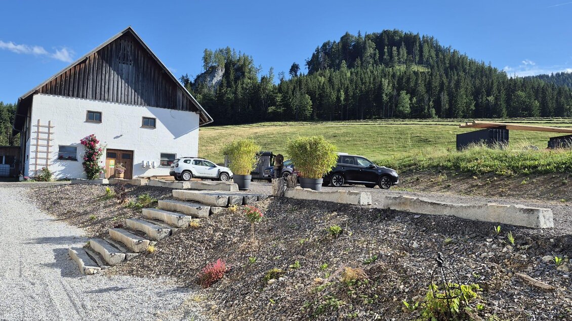 Ein Bauernhaus mit einem gepflasterten Vorplatz und mehreren Autos. Im Hintergrund sind grünen Wiesen und bewaldete Hügel zu sehen. | © TV Hochsteiermark