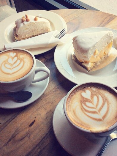 Zwei Tassen Kaffee mit kunstvollem Milchschaum und zwei Stück Kuchen auf einem Holztisch. Es ist eine gemütliche Café-Atmosphäre. | © Shutterstock / Mutita Narkmuang