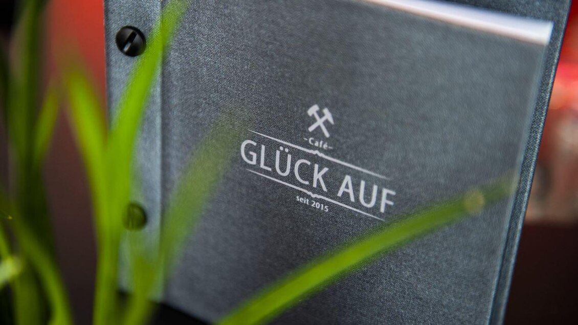 Ein schönes Café-Menü mit dem Titel "GLÜCK AUF" und dem Gründungsjahr 2015. Im Hintergrund ist ein grünes Pflanze zu sehen. | © Pfripfl Lucas